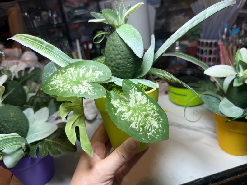 Op de afbeelding: Een fel limoengroene keramische pot met een kunstplantarrangement. Het display bevat een centraal, getextureerd, donkergroen, fruitachtig element omringd door bonte, hartvormige groen-witte bladeren en lang, slank blad.