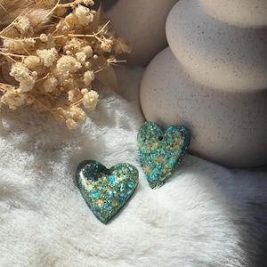 May include: Two heart-shaped pendants crafted from resin, featuring a deep green base embedded with a dense mixture of iridescent teal, gold, and emerald glitter particles that catch the light, creating a textured and reflective surface finish.