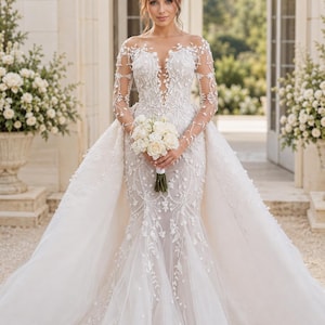 Puede incluir: Un vestido blanco largo hasta el suelo con mangas largas transparentes, un escote ilusión profundo y un intrincado bordado de encaje floral. El vestido tiene un corpiño ajustado que se ensancha en una voluminosa sobrefalda de tul, con un ramo de rosas blancas.