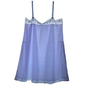 May include: A light purple sleeveless chemise featuring thin shoulder straps, a V-neckline, and a hemline trimmed with intricate white floral lace detailing. The garment has a loose, A-line silhouette with small side slits and matching lace trim.