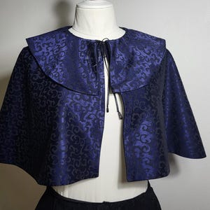 May include: A dark blue cape with a floral pattern. The cape has a wide collar and a tie closure at the neck. The fabric has a subtle sheen. The garment is displayed on a mannequin.