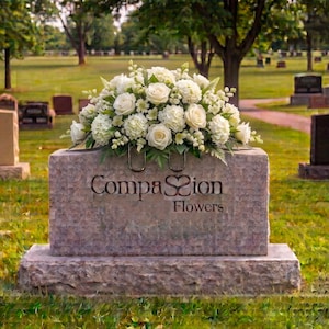 May include: A large floral arrangement of white roses and hydrangeas sits atop a rectangular pink granite headstone. The stone features the engraved text Compassion Flowers with a heart-shaped symbol replacing the letter s in Compassion.