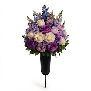 May include: A floral arrangement featuring purple, lavender, and cream roses mixed with blue delphinium and green foliage. The flowers are contained in a black, tapered, spike-bottomed vase designed for insertion into the ground.