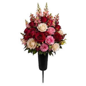 May include: A floral arrangement featuring deep red, light pink, and cream-coloured roses mixed with tall, slender pink snapdragon stalks. The flowers are densely packed and set into a plain, matte black cylindrical vase with a pointed ground stake.
