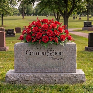 May include: A rectangular grey stone monument featuring the text Compassion Flowers with a heart-shaped symbol replacing the double s. A large, dense arrangement of vibrant red roses and small red filler flowers sits atop the stone surface.