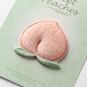 Puede incluir: Un parche bordado con forma de melocotón con textura de felpa rosa claro y dos pequeñas hojas verdes en la base. El parche está sujeto a una tarjeta de color verde claro que presenta el texto Sweet Peaches y una frase más larga abajo.