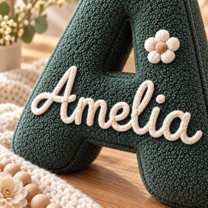 May include: A dark green, textured, plush letter A cushion featuring the name Amelia embroidered in white cursive script across the front. A small, cream-coloured, five-petalled fabric flower is attached to the upper right side of the letter.