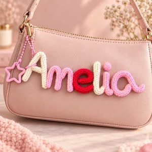 May include: A light pink shoulder bag featuring a custom name charm that spells Amelia in knitted yarn letters. The letters alternate between cream, light pink, and red colors. A small pink star-shaped charm hangs from a beaded chain on the side.