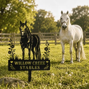 May include: A black metal garden sign featuring a silhouette of a horse standing between two floral stems. The rectangular base displays the text "WILLOW CREEK STABLES" in a serif font, accented by small heart shapes and decorative leaf motifs.