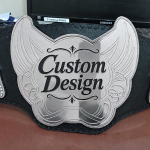 Könnte beinhalten: Ein Meisterschaftsgürtel mit einer großen, polierten silberfarbenen Metall-Mittelplatte mit verzierten flügelartigen Seitenteilen. Die Platte zeigt den schwarzen Text Custom Design in einer fetten Serifenschrift auf einem schwarzen Lederriemen.