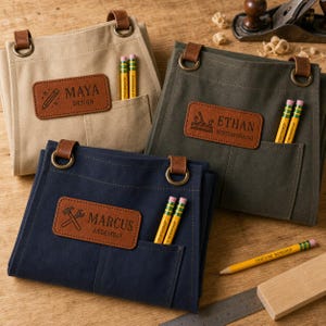 May include: Three canvas waist aprons in beige, olive green, and navy blue. Each features a brown leather patch with a name and icon, two metal D-rings, and a front pocket holding two yellow pencils. The patches read MAYA DESIGN, ETHAN WOODWORKING, and MARCUS ASSEMBLY.