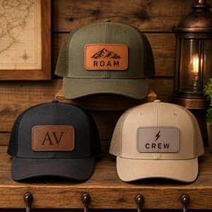 May include: Three mesh-back baseball caps in olive green, black, and tan. The olive cap features a tan leather patch with a mountain graphic and the text ROAM. The black cap has a brown leather patch with the letters AV. The tan cap has a grey leather patch with a lightning bolt icon and the text CREW.