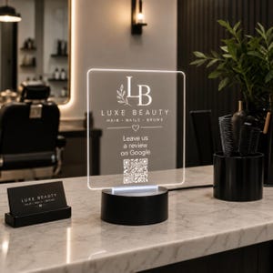 May include: A clear acrylic sign with a white LED light base featuring the Luxe Beauty logo, the text Leave us a review on Google, and a QR code. Next to it sits a black business card holder with a matching Luxe Beauty card and a black cup.