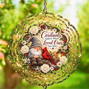 May include: A circular metal wind spinner featuring a gnome with a grey hat and a red cardinal bird. The design includes a floral wreath with white roses, daisies, and a lantern. The text reads: Cardinals Appear When Loved Ones Are Near.