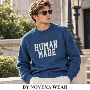 Könnte beinhalten: Ein blauer, langärmeliger Strickpullover mit dem Schriftzug HUMAN MADE in weißen Blockbuchstaben auf der Brust. Der Pullover hat einen gerippten Rundhalsausschnitt, gerippte Bündchen und einen gerippten Saum im klassischen Raglan-Design.