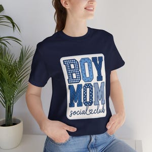Könnte beinhalten: Ein marineblaues Kurzarm-T-Shirt mit einer großen rechteckigen Grafik, die den Text "BOY MOM" in Blockbuchstaben zeigt, gefüllt mit verschiedenen blau-weißen Mustern, gefolgt von dem kursiven Text "social club" mit einem Herzsymbol.