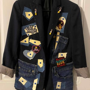 Op de afbeelding: Een zwarte blazer met denim patches, gele stoffen accenten en blauwe studs. Het kledingstuk bevat geborduurde patches met de tekst 'a lil bougie', 'Black princess' en 'BLACK GIRLS BEEN MAGIC', samen met versieringen van veiligheidsspelden.