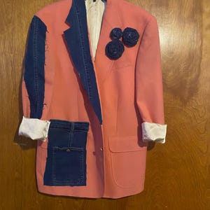 May include: A salmon-colored blazer featuring a dark blue denim lapel, a denim pocket patch on the lower front, and three dark blue fabric flower embellishments on the upper chest. The sleeves are cuffed to reveal a white lining.