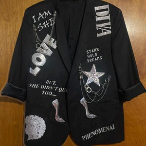 May include: A black blazer featuring silver sequinned embellishments, including the words I AM SHE, DIVA, LOVE, PHENOMENAL, and STARS HOLD DREAMS. It includes silver chain accents, a sequinned star, two high-heeled shoe motifs, and a textured circular patch.
