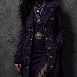 May include: A long, deep purple suede-textured coat with silver-toned buttons, worn over a matching ribbed turtleneck dress. The outfit includes a wide belt with a large silver buckle, layered silver chain necklaces, and black lace patterned tights.