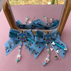 May include: Two light blue hair bows featuring a repeating cat pattern, a central cat face charm, and dangling beaded strands spelling DERPY. Each bow includes a metallic teal ribbon accent and a silver-toned charm branch with heart-shaped leaves.