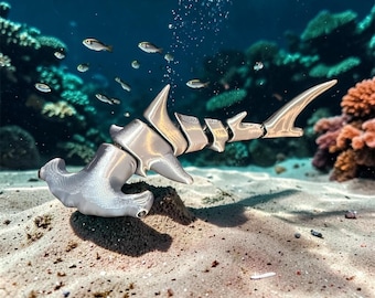 May include: A silver-toned, articulated hammerhead shark figurine with a segmented body design. The shark is positioned on a sandy seabed, featuring a metallic finish with visible texture on the head and smooth, curved fins along its body.