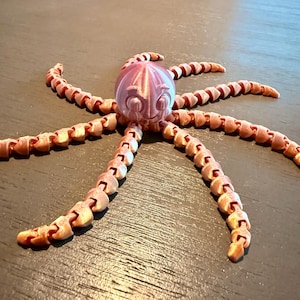May include: A 3D printed articulated octopus figurine with a shiny, metallic light pink finish. The creature features a round head with large, expressive eyes and eight long, segmented, flexible tentacles that extend outward in a radial pattern.