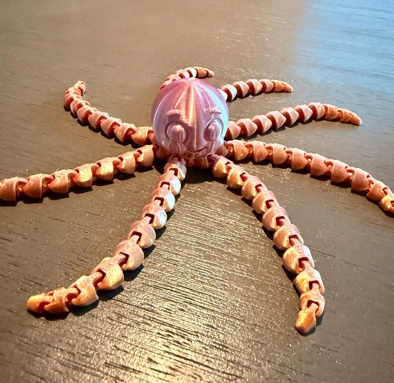 May include: A 3D printed articulated octopus figurine with a shiny, metallic light pink finish. The creature features a round head with large, expressive eyes and eight long, segmented, flexible tentacles that extend outward in a radial pattern.