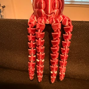 May include: A bright red, 3D-printed octopus figurine with a smooth, rounded head featuring embossed eye details and a mouth. The creature has four long, articulated, segmented tentacles that hang vertically, showing a layered plastic texture.