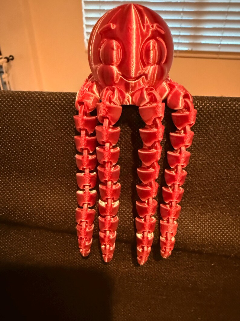 May include: A bright red, 3D-printed octopus figurine with a smooth, rounded head featuring embossed eye details and a mouth. The creature has four long, articulated, segmented tentacles that hang vertically, showing a layered plastic texture.