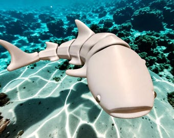 May include: A light beige, articulated shark figurine with a segmented body design. The shark features a smooth, matte surface, a rounded head, visible side fins, and a tail fin, captured in a swimming pose against a bright underwater background.