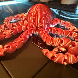 May include: A 3D printed red octopus figurine with a smooth, rounded head featuring embossed eyes and a smiling mouth. The creature has eight articulated, segmented tentacles that extend outward, showcasing a shiny, metallic-like finish.
