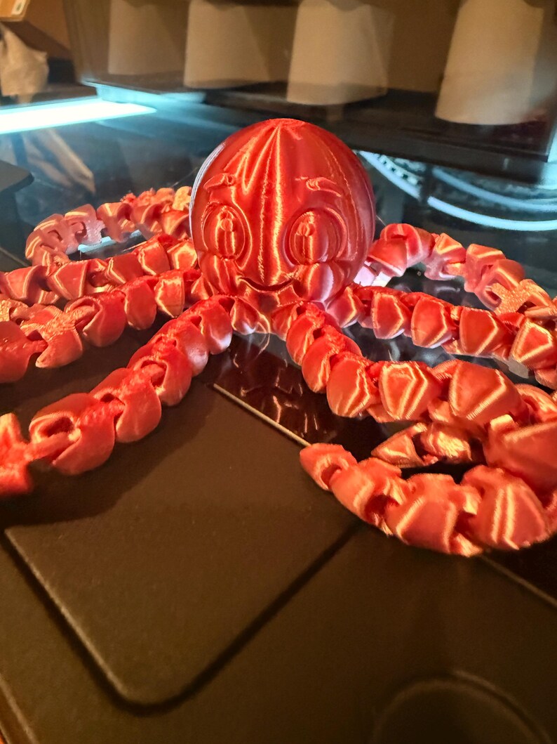May include: A 3D printed red octopus figurine with a smooth, rounded head featuring embossed eyes and a smiling mouth. The creature has eight articulated, segmented tentacles that extend outward, showcasing a shiny, metallic-like finish.