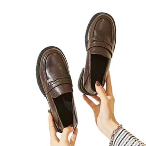 May include: A pair of dark brown leather penny loafers featuring a rounded toe, a classic saddle strap across the vamp, and a thick, dark-coloured rubber sole. The shoes have a smooth, polished finish and a structured silhouette for everyday wear.