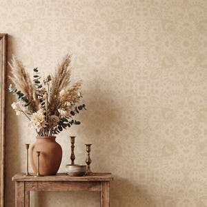 May include: A beige wallpaper featuring a repeating mandala-inspired pattern in a lighter shade. The wall is decorated with a terracotta vase filled with dried pampas grass and flowers, placed on a wooden table with brass candlesticks and a small bowl.