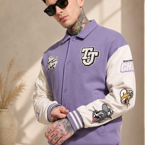 May include: A light purple varsity jacket with cream-colored sleeves, featuring embroidered patches of Tom and Jerry characters, the text "TJ" on the chest, and "The Cat & Mouse" on the sleeve, finished with striped ribbed cuffs and a snap-button front.