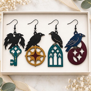 May include: A set of four drop earrings featuring black raven silhouettes. The first shows a raven on a teal key, the second a raven on a gold star wheel, the third a raven on a teal arched window, and the fourth a blue raven on a maroon crescent.