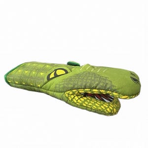 May include: A green oven mitt shaped like an alligator head, featuring a textured scale pattern, yellow eyes, and an open mouth revealing white teeth and a red interior. The mitt is constructed from fabric and designed for kitchen use.