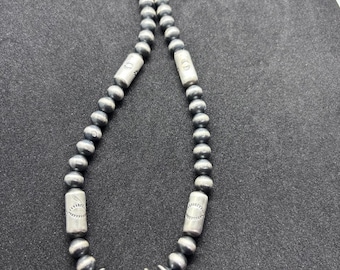 May include: A silver-toned necklace featuring a series of round, polished beads interspersed with four cylindrical metal beads. Each cylindrical bead is engraved with a distinct eye-shaped motif. The necklace ends with several pointed, biconical beads.