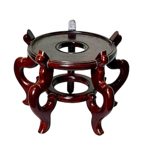 May include: A dark reddish-brown wooden display stand featuring a circular top platform with a central opening, supported by four curved, ornate legs that connect to a smaller circular base ring near the bottom, creating a stable elevated structure.