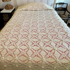 Pode incluir: Uma colcha de crochê na cor creme com um padrão de diamante repetitivo e pequenos detalhes florais em relevo em cada interseção. O tecido intrincado e aberto está estendido sobre uma cama com uma cabeceira de metal de cor clara.