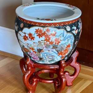 May include: A round ceramic fishbowl planter featuring a black background with gold speckles, decorated with colorful hand-painted birds, red maple leaves, and floral motifs. The bowl rests on a dark reddish-brown carved wooden display stand.