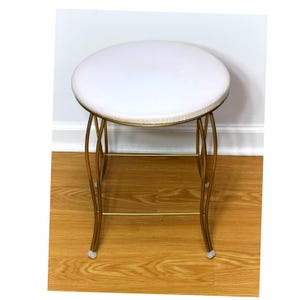 May include: A round stool featuring a white padded seat cushion and a gold-toned metal frame. The base consists of four curved legs connected by horizontal support bars, with small protective caps on the feet of each leg.