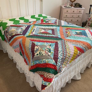 May include: A colorful patchwork quilt with a white lace bed skirt. The quilt is made of various fabrics in shades of red, blue, green, and yellow. The quilt is on a white bed frame with a green and white patterned bedspread.