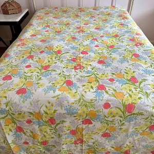 Puede incluir: Una colcha floral blanca con un patrón repetido de tulipanes rosas, flores amarillas y flores azules. La colcha está sobre una estructura de cama de metal blanco. El diseño general es un patrón floral vintage.