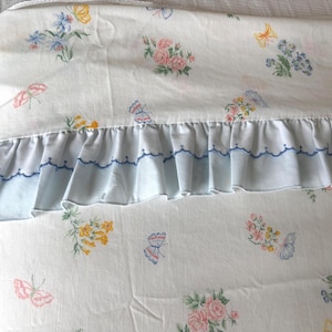 May include: A white bed sheet featuring a repeating pattern of pink roses, blue flowers, and yellow and pink butterflies. The sheet includes a light blue ruffled edge with a decorative dark blue scalloped embroidery stitch along the seam.