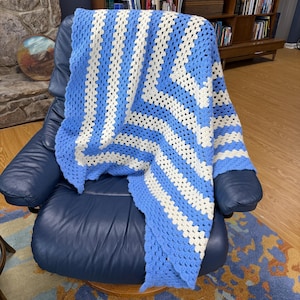 May include: A handmade crochet blanket in shades of light blue and white, draped over a blue leather armchair. The blanket features a striped pattern, with alternating bands of blue and white. The crochet stitches are clearly visible, showcasing the craftsmanship.