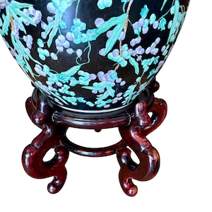 May include: A large, rounded ceramic vase with a glossy black background featuring a painted pattern of light green leaves and clusters of purple berries. The vase rests on a dark reddish-brown wooden stand with four curved, ornate legs.