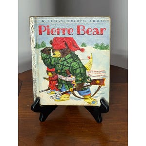 May include: A Little Golden Book titled "Pierre Bear" featuring a bear character in a red hat and green plaid jacket, holding a rifle. The cover art depicts a snowy scene with two bears. The book is displayed on a black stand.