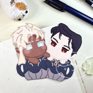 May include: A pair of acrylic charms featuring two chibi-style characters with dark skin and light hair, and light skin and dark hair, both wearing dark blue scalloped-edge outfits with gold accents, positioned together on a white surface.